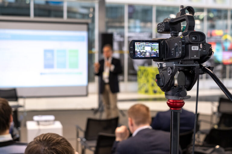 LiFi Media Production Corporate Video Trends 2025 Video camera on tripod recording presentation of male coach standing on stage with display in front of audience