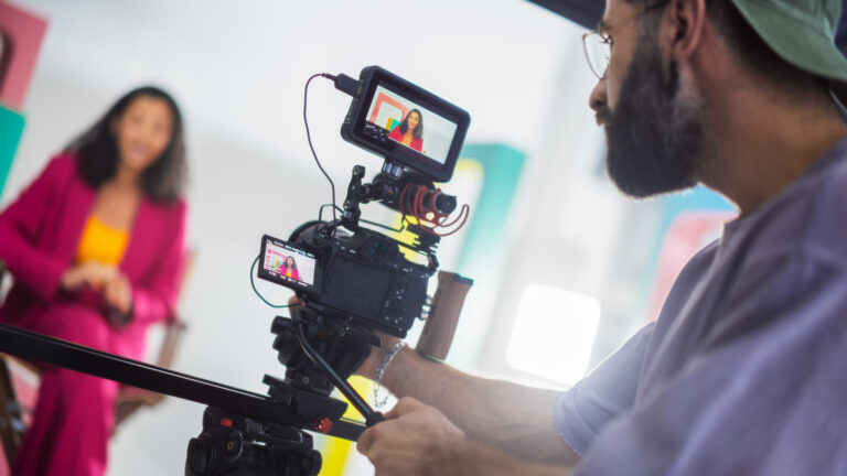 LiFi Media Production Brand Videography, film set with a young black female host in a pink suit engaging in a lively discussion captured by a male cinematographer focusing intently on his camera equipment surrounded by colorful backdrops